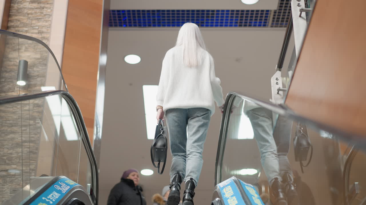 back view of browsing shopper riding moving walkway in mall interior carrying handbag gliding upward past glass railing into busy retail environment with nearby shoppers moving behind