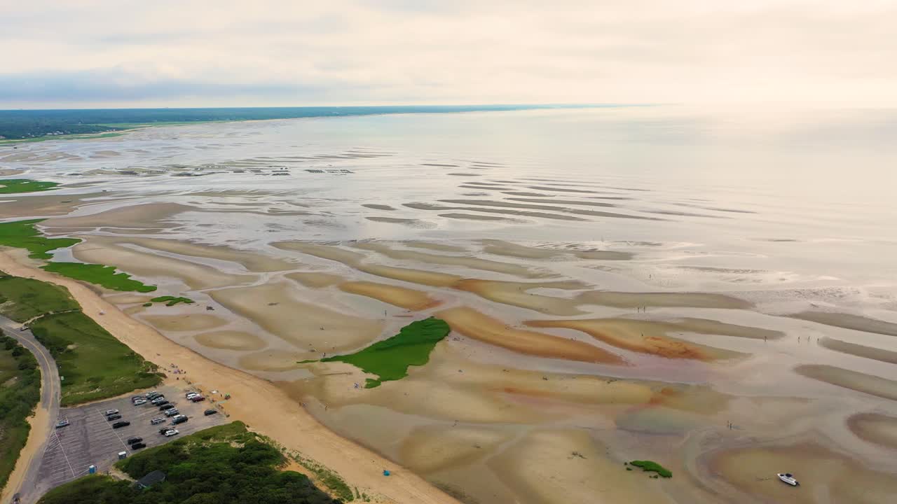 Sweeping drone video shows tidal flats stretching to the horizon, with tide pools and ridges reflecting light, while a neighborhood of homes frames a lively public beach along the coast