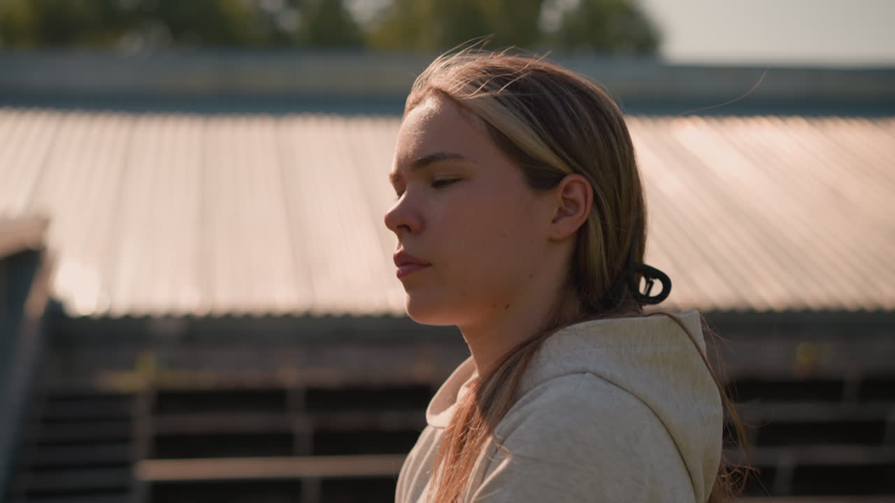 머리카락이 뒤로 여있는 재색 후디를 입은 젊은 여성은 빛이 그녀의 얼굴에서 반사되어 따뜻한 빛을 내며, 배경은 흐릿한 빈 경기장 좌석을 보여줍니다.