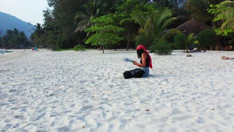 Trendy-girls-with-red-head-scarf-and-travel-bag-left-on-white-sand-of-quiet-exotic-beach,-reading-tourist-guide-of-tropical-island-in-Vietnam