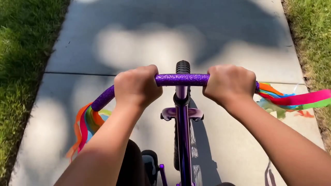 Top-down point-of-view shot showing a child’s hands on glittery purple handlebars as they ride a small purple bike with rainbow streamers along a sunlit