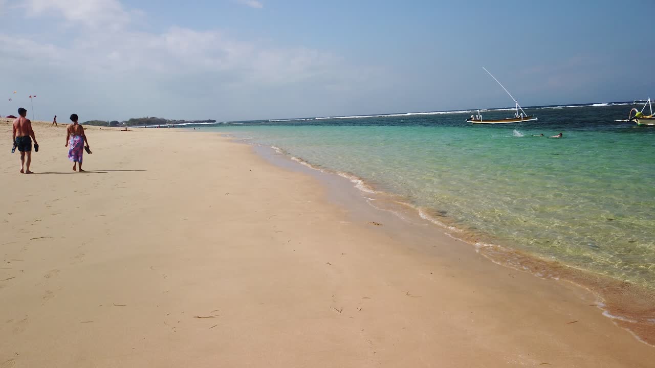 Nusa Dua beach, beautiful summer day and exotic destination. Peoples walking around beach on white sand with amazing clear blue ocean in background
