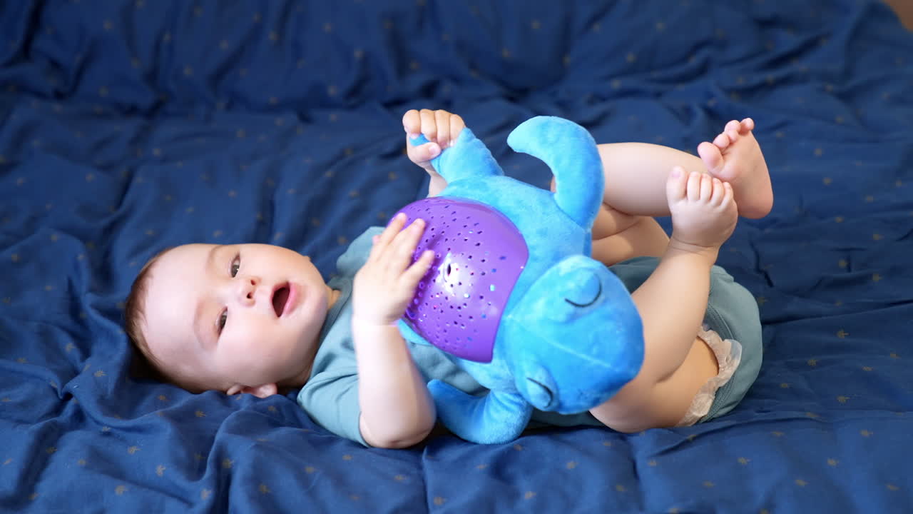 Cute Caucasian toddler lies on the soft bed on his back. Baby boy holding a big toy turtle on his belly playing with it. Side view.