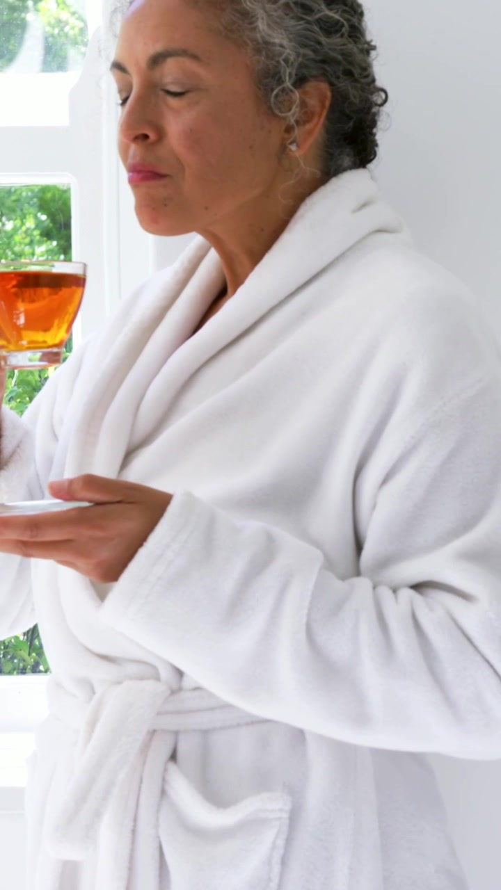 Vertical video: Sipping woman in white bathrobe by window, savoring tea aroma from glass teacup
