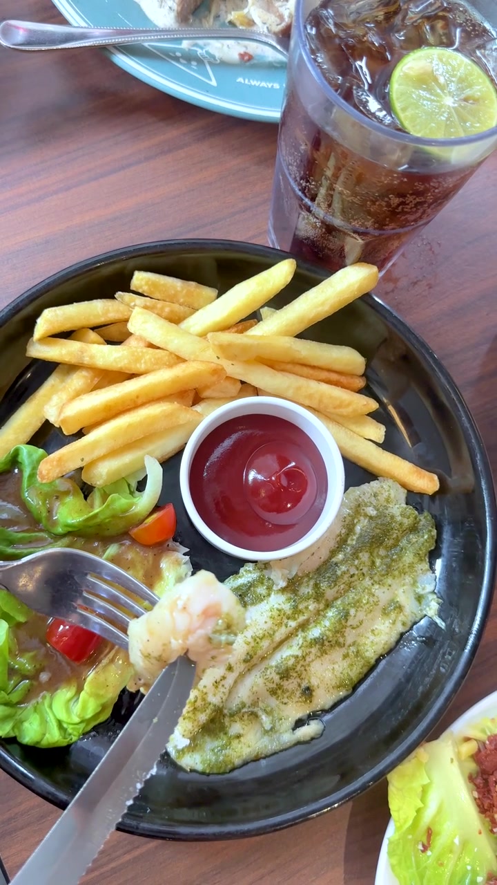Inquadratura dall'alto di una persona che taglia pesce alla griglia servito con patatine fritte, insalata e cola
