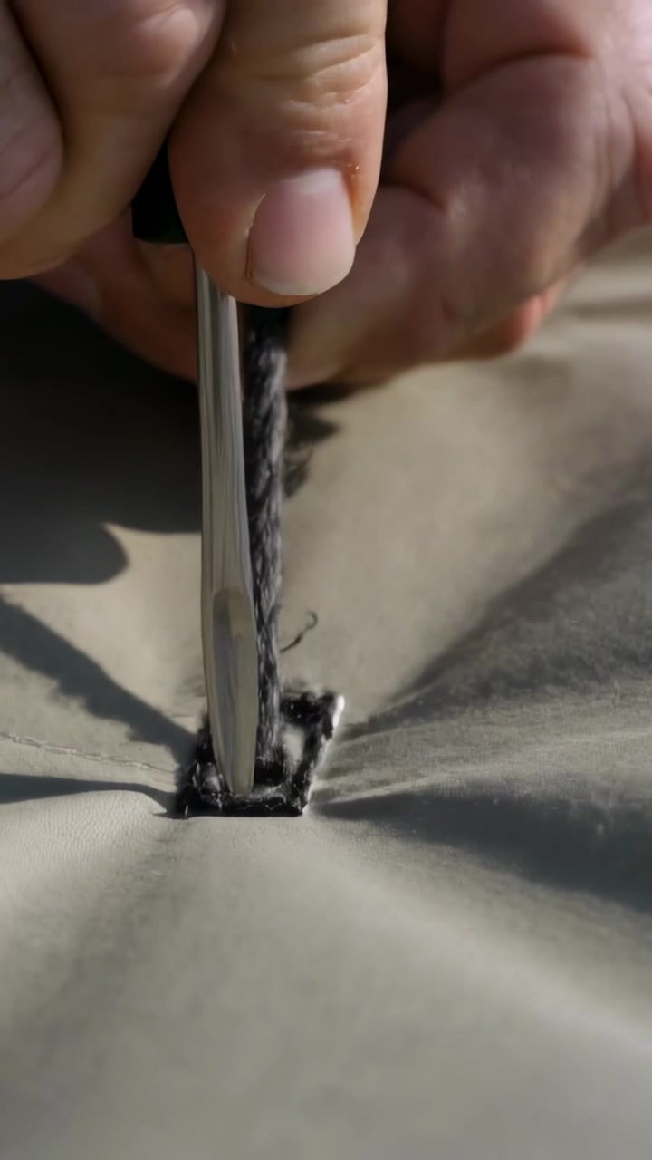 Video verticale: La punta di un attrezzo entra in un occhiello, mani liberano un cordino sfilacciato su tessuto color kaki con delle pinze.