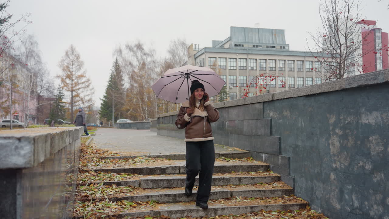 Smiling lady making phone call while holding umbrella, wearing black knit cap, brown shearling jacket, black trousers, walking down stone steps during light snowfall with colorful autumn leaves