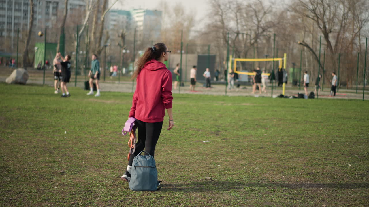 Individual With Backpack Strolling Through Park, Person In Casual Attire Passing Sports Field In Park, Observer With Backpack Leisurely Walking Past Playing Field Situated In Suburban Park Area