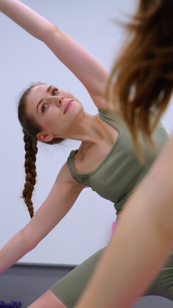 primer plano de una dama con una camiseta marrón participando en ejercicios de fitness estirándose con una cinta en un gimnasio, otra dama con un traje verde fluido cerca, equipos dispuestos en una mesa