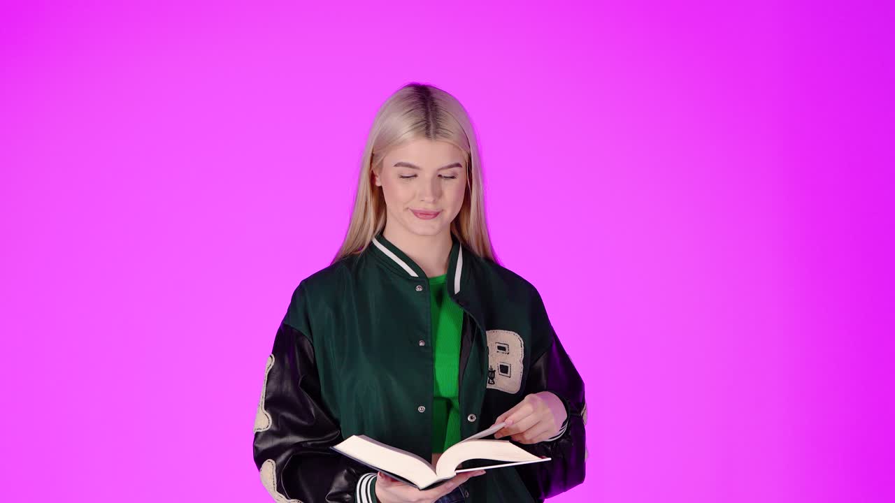 Giovane studentessa bionda che si gode la lettura di un libro e sorride, ritratto in studio con sfondo rosa.