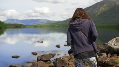 Chica-Caminando-Por-La-Orilla-De-Un-Hermoso-Lago-Con-Un-Claro-Reflejo-Perfecto-De-Las-Montañas-Alrededor