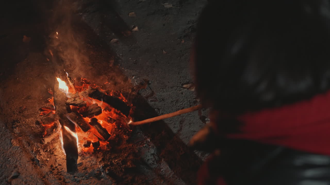 Pemandangan kemping malam dengan api unggun, seorang pendaki bersyal merah mengipasi bara api yang menyala dengan tongkat sambil memanggang marshmallow, jaket kulit dan tangan terlihat, abu dan percikan api membumbung ke udara berasap, suasana akrab.