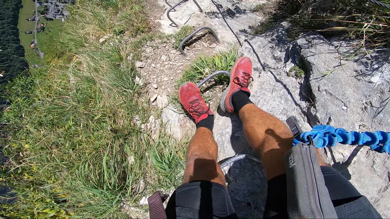 POV action camera shot of a man taking steps down a very steep cliff on the Via Ferrata climbing route in the Swiss alps during a hot day with bright sunlight. Wide angle facing down.