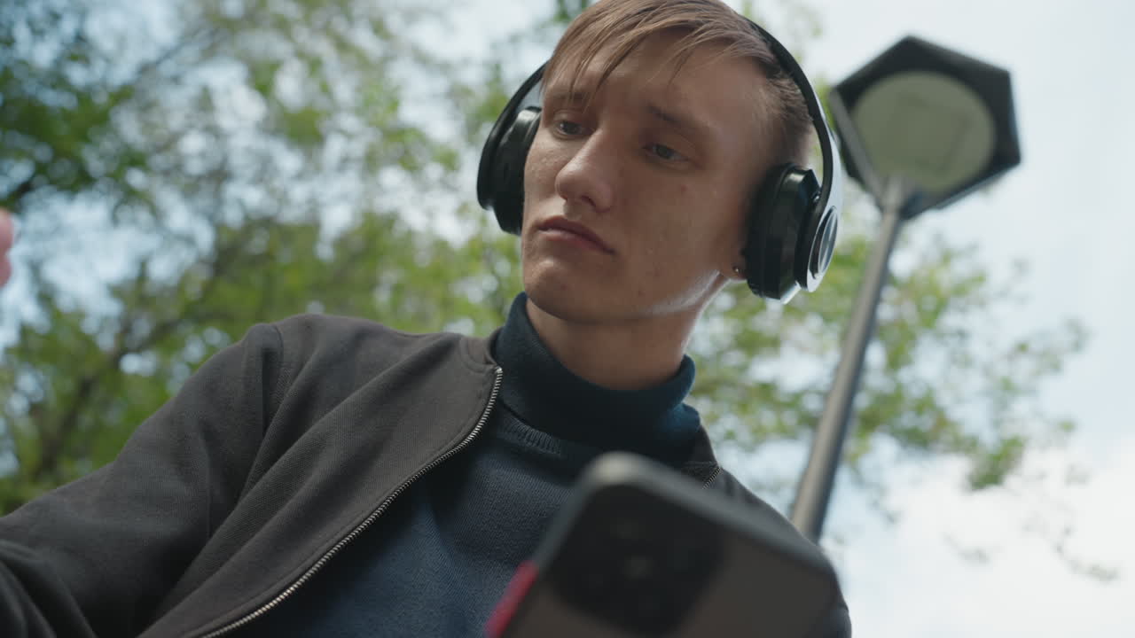 Giovane uomo caucasico che controlla il telefono con le cuffie, primo piano dal basso che mostra lo schermo dello smartphone e il lampione, alberi del parco sullo sfondo, scorrendo tra le app con espressione attenta, moderno