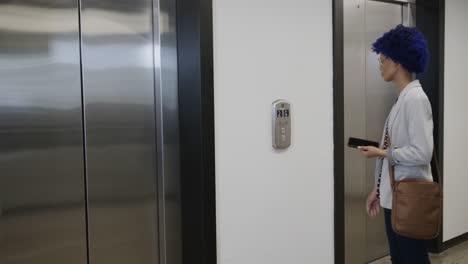 mujer de negocios biracial con afro azul usando un teléfono inteligente y entrando en el ascensor en la oficina, cámara lenta