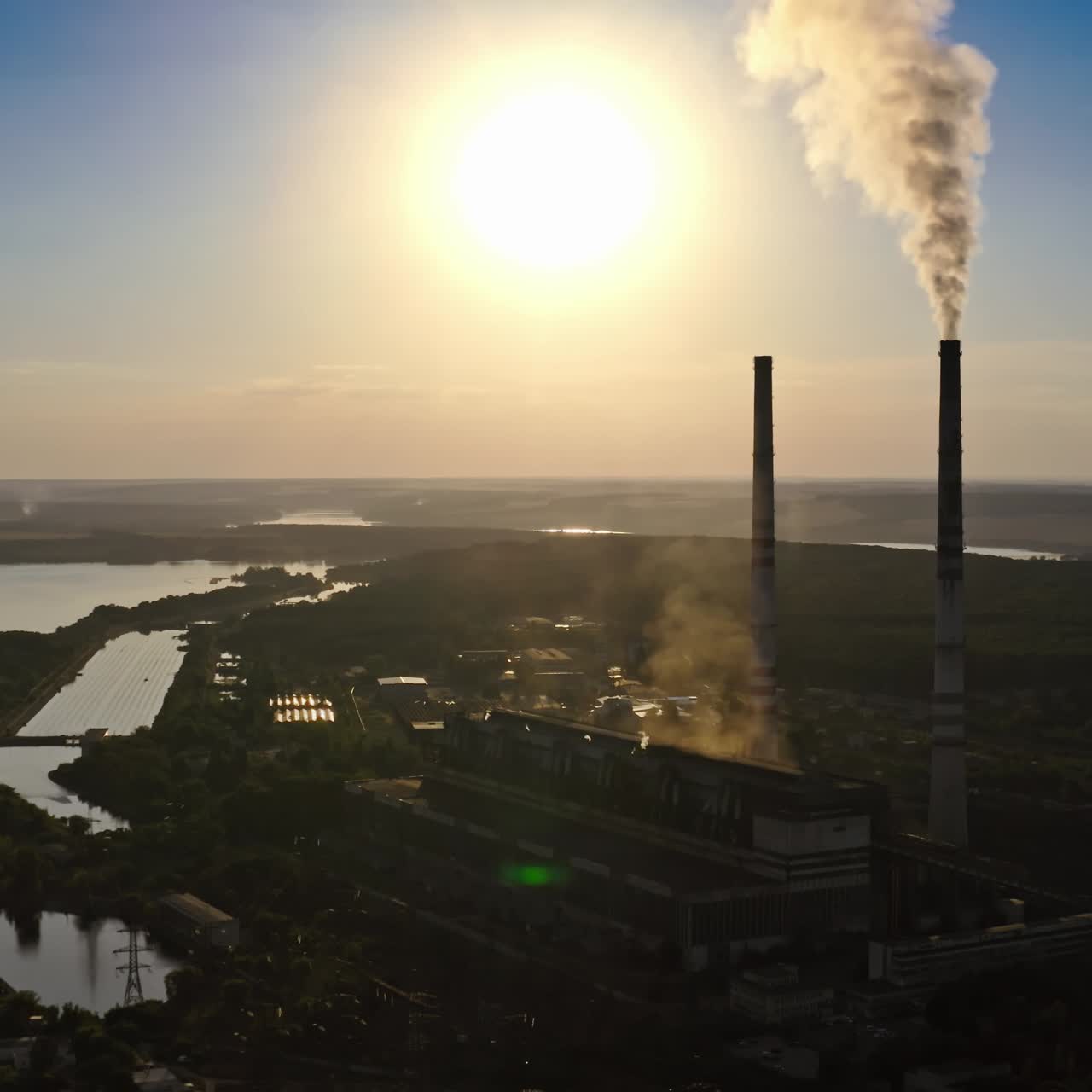 Dark factory against the orange setting sun. Factory pipes with smoke. Dirty smoke pours out of chimneys into the air on the river background. Aerial view video