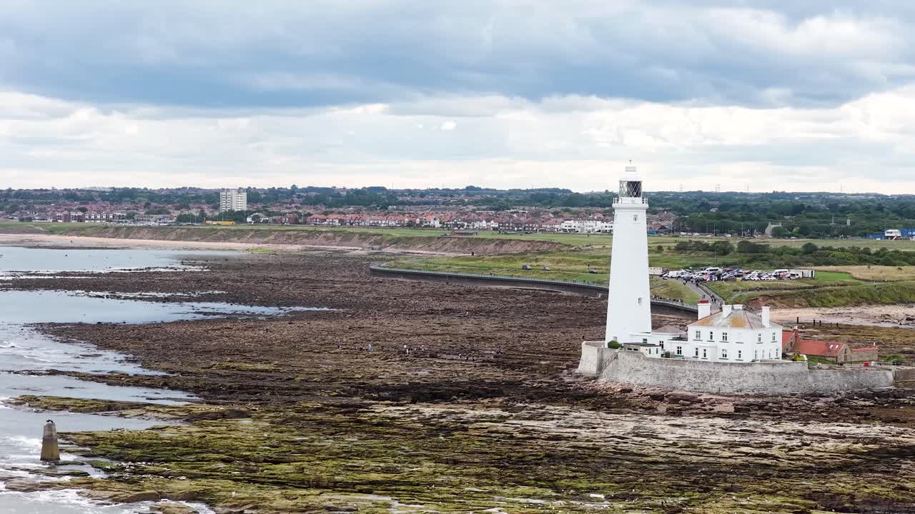 Aerial camera smoothly pans across St Mary’s Lighthouse and surrounding rocky shoreline under overcast daylight, revealing coastal landscape and distant townscape in Whitley Bay, England