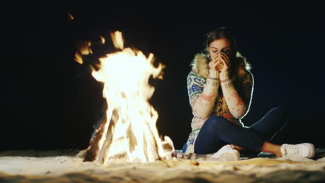 a young woman sitting alone by the fire drinking tea looking at the fire