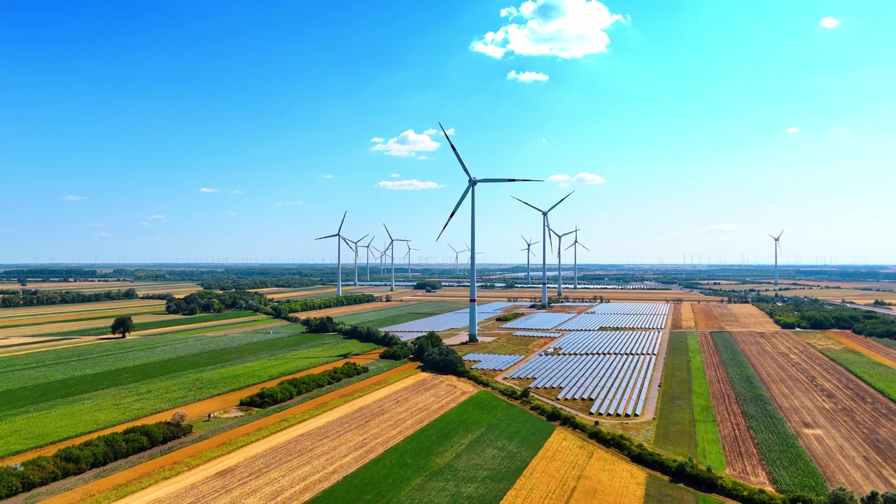 Wind turbines and solar panels in agricultural fields. Aerial view of wind turbines and solar panel arrays among green and yellow agricultural fields under blue sky.
