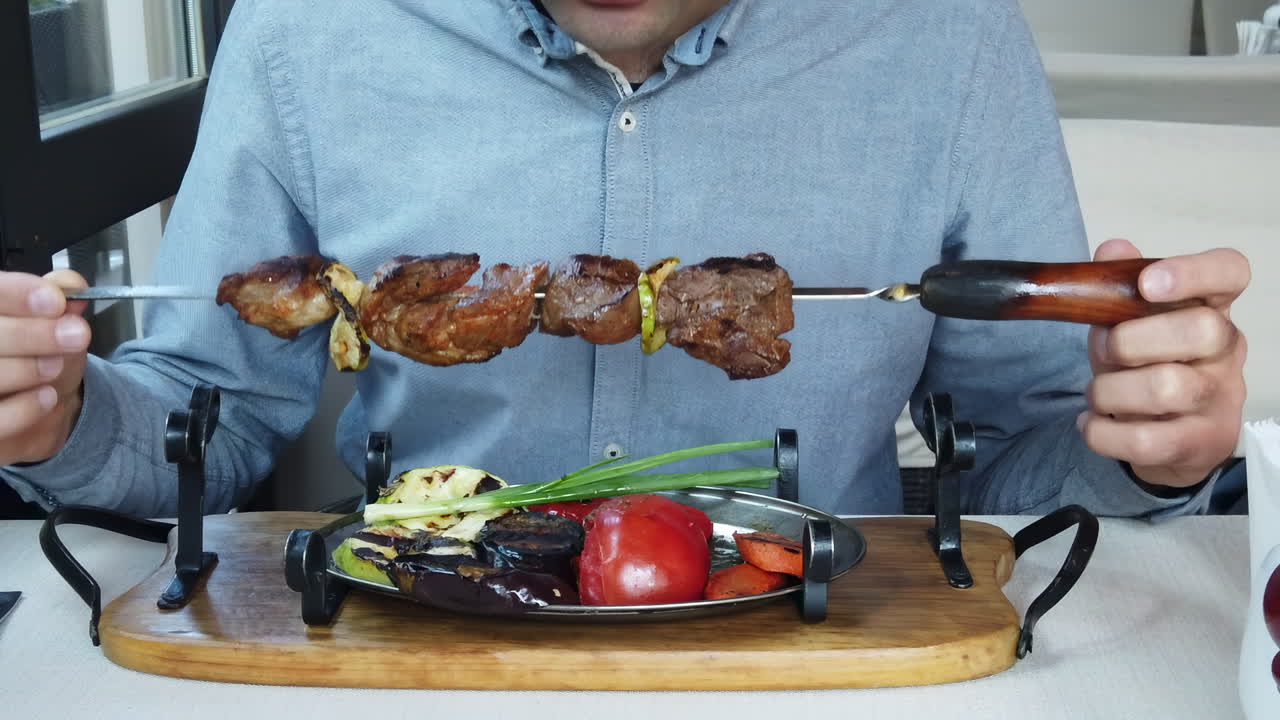 Close up of a man holding a skewer of grilled meat with roasted vegetables on a plate