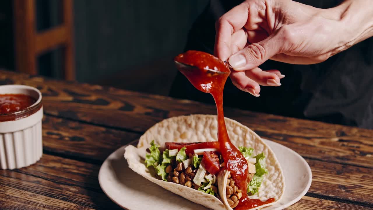 Close-up angle of a hand adding sauce to a taco on a wooden table, capturing a rustic and appetizing
