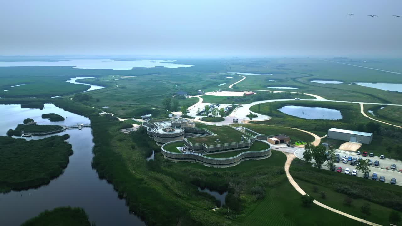 Hazy summer aerial descends and circles eco centre amid sprawling wetlands
