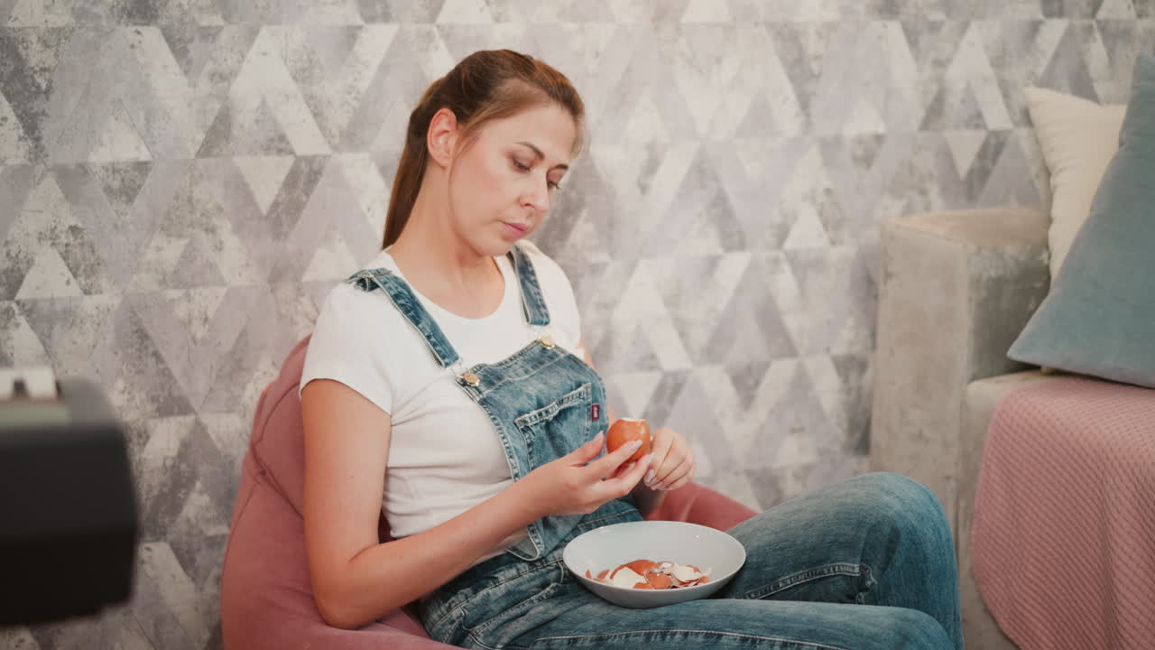 Relaxed woman in white shirt and denim overalls seated on pink beanbag peeling egg shell into bowl on lap, with soft furnishings and partial view of digital piano and couch in cozy indoor setting