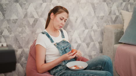 relaxed woman in white shirt and denim overalls seated on pink beanbag peeling egg shell into bowl on lap, with soft furnishings and partial view of digital piano and couch in cozy indoor setting