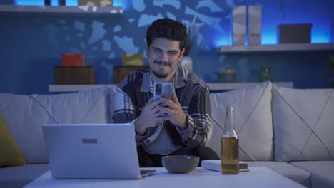 un joven feliz enviando mensajes de texto por teléfono por la noche. hay alcohol en la mesa.
