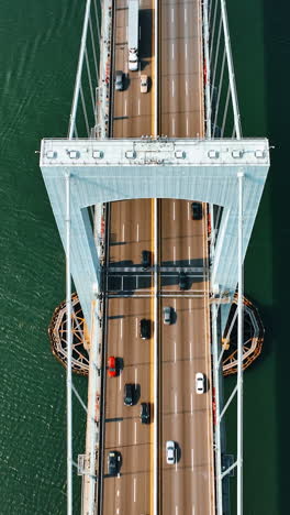 part of the suspension bridge over sea-green water. cars move by the roads on the bridge under the support. motor boat is on the water. top view. vertical video