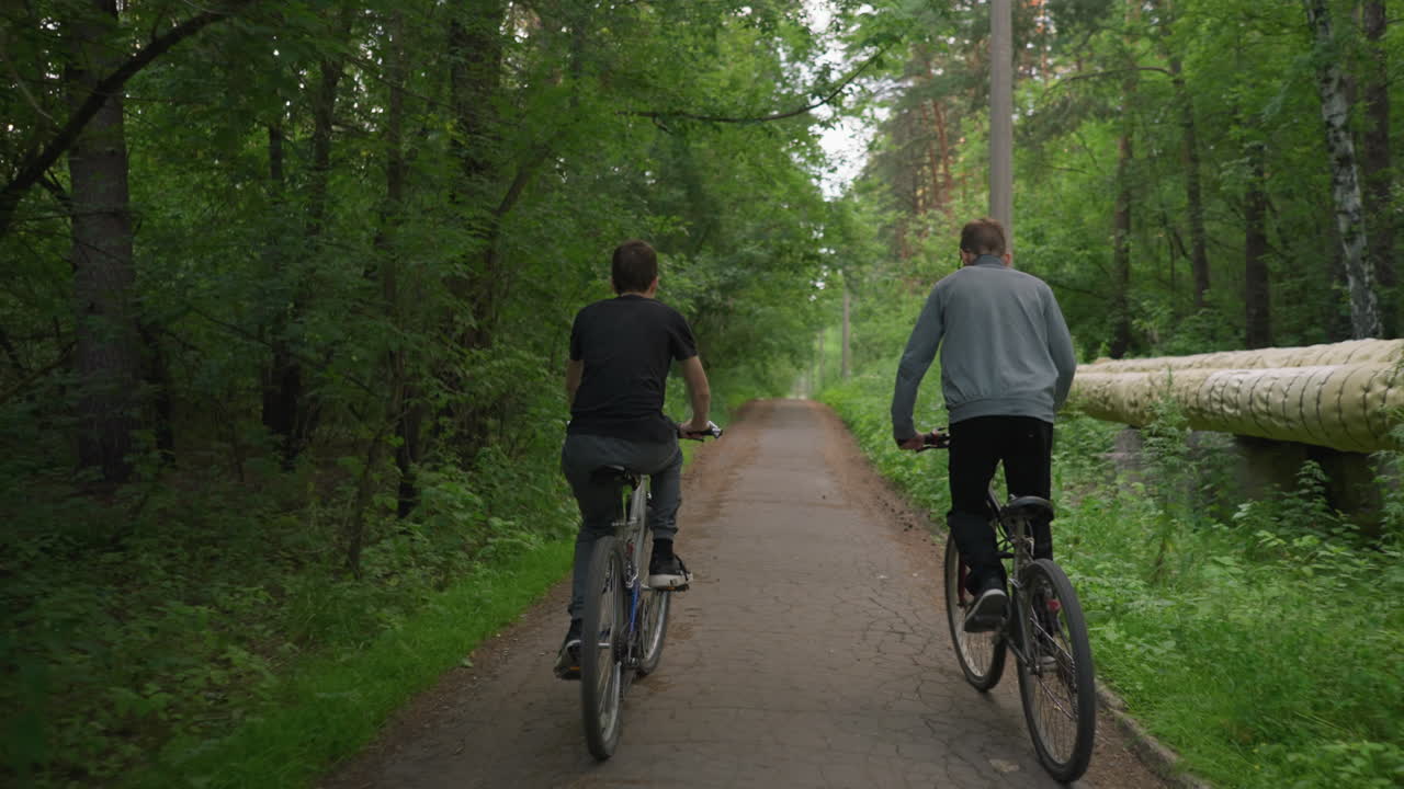 두 친구가 초록색 나무로 둘러싸인 포장된 길을 따라 자전거를 타고 자연 속에서 느하게 타는 것을 즐기고, 회색 을 입은 사람이 타는 동안 서 있습니다.