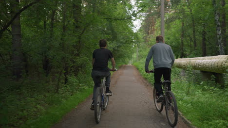 two friends riding bicycles along a paved path surrounded by lush green trees, enjoying a leisurely ride amidst nature, with the one in grey top standing while riding