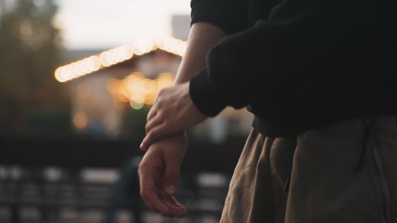 Primo piano di mani caucasiche che rimboccano una manica contro luci bokeh. Lo sfondo crepuscolare della città con lampadine calde incornicia la scena, mostrando le mani che si protendono, toccando il polso, una pausa consapevole prima del turno di notte, sia per un artista che per un pendolare.