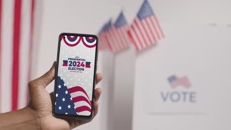 Close-Up-Of-Hand-Holding-Mobile-Phone-With-Screen-Encouraging-People-To-Vote-In-2024-US-Presidential-Election