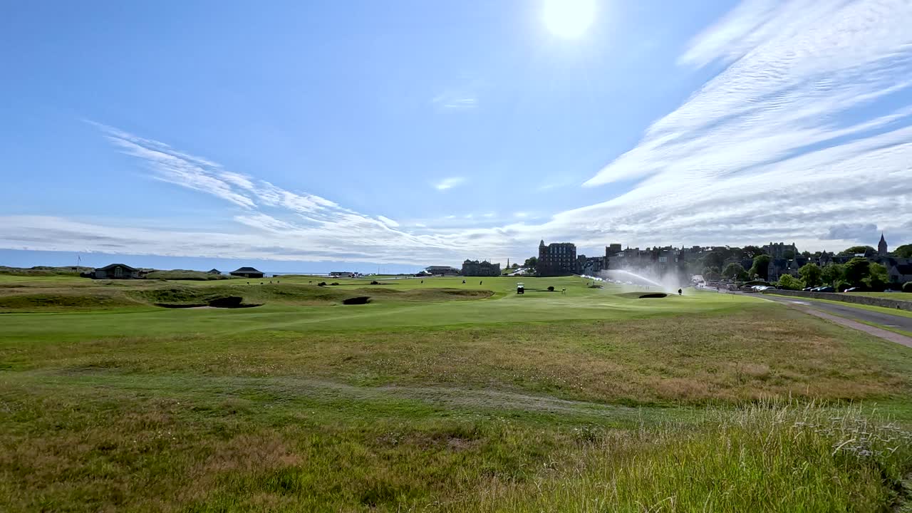 Wide shot of green golf course, bright sunlight, blue sky, sprinklers watering grass, static camera