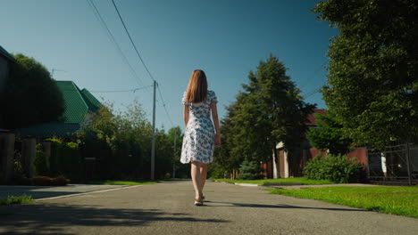 back view of fair skin woman in floral dress walking alone down sunny suburban road surrounded by green trees, power lines, and residential houses with calm morning light and peaceful atmosphere