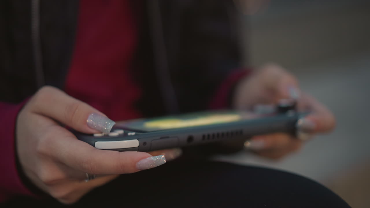 CloseUp Hands Holding Handheld Console In Dim Urban Light, Manicured Nails On Controller Grips, Thumb On Joystick, Casual Jacket Sleeve, Cozy Focus On Device Interaction And Idle Screen