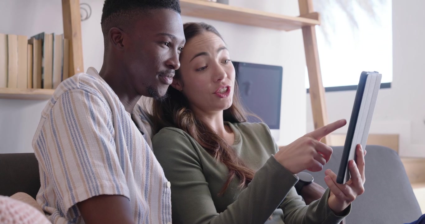 Pareja diversa acurrucada en el sofá compartiendo una tablet, señalando, deslizando y riendo con el contenido.