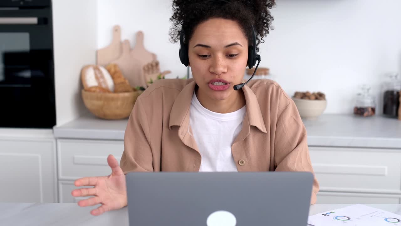 Positive successful african american curly-haired girl, in casual clothes, working or studying from home, using laptop and headset, talking on video conference, smiling friendly