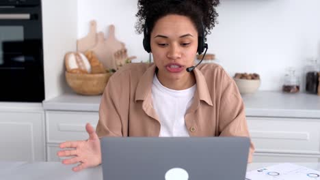 exitosa chica afroamericana de cabello rizado, con ropa casual, trabajando o estudiando desde casa, usando portátil y auriculares, hablando en videoconferencia, sonriendo amistosamente