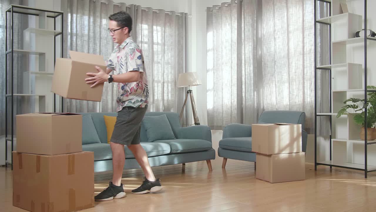 아시안 남자가 휴식을 취하기 위해 소파에 앉기 전에 새로운 집으로 물건과 함께 카드보드 상자를 운반합니다. 그녀는 새로운 집으로 이사합니다.