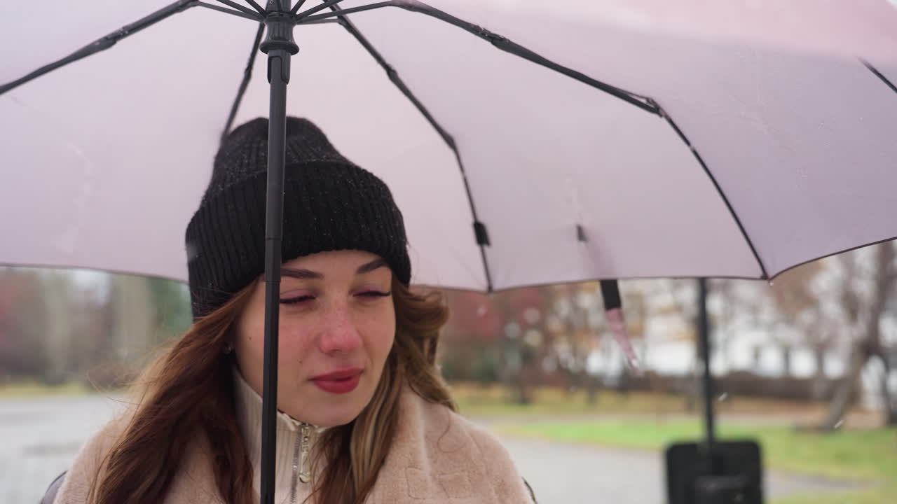 Cheerful girl holding umbrella, wearing black knit cap and brown shearling jacket, walking calmly on cold day with light snowfall, surrounded by peaceful outdoor urban landscape