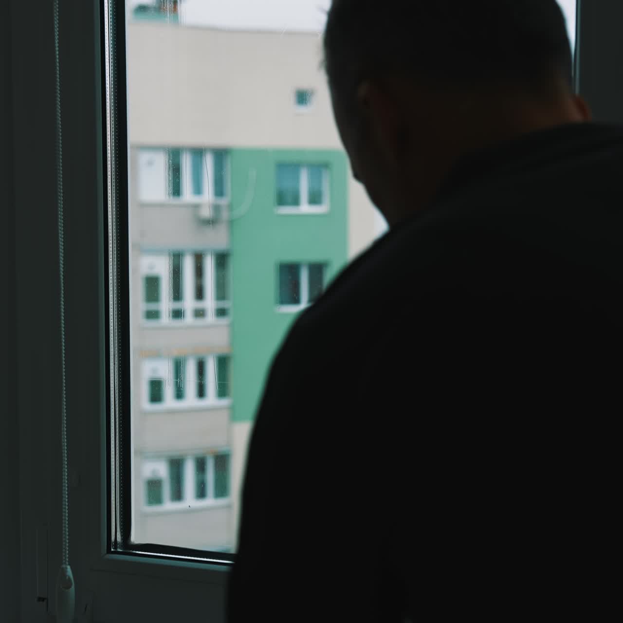 Man cleans the window and squeezes water with a brush. Male worker washing glass inside the flat. Block of flats background through clean window. Householding chores