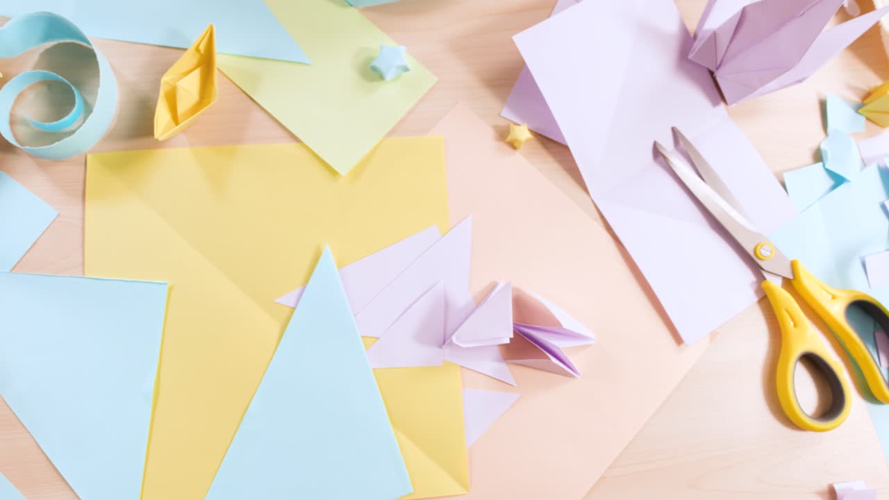 Una lenta panoramica della camera rivela carta color pastello, forbici e origami a forma di gru piegati su un tavolo