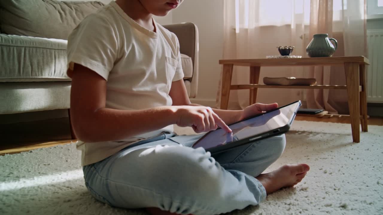 Bambino in età scolare in maglietta e jeans, che tocca il tablet in grembo in salotto, giocando a un gioco.