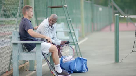 Happy-diverse-male-friends-sitting-on-bench-talking-at-tennis-court,-slow-motion