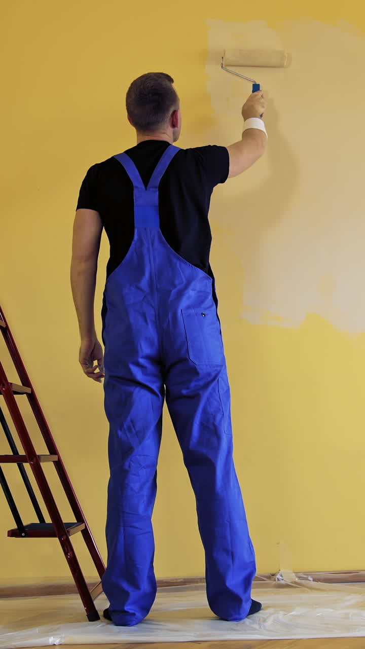 Room decoration with new color. Professional worker in blue overalls painting wall with a paint roller in empty room. Rear view. Vertical video