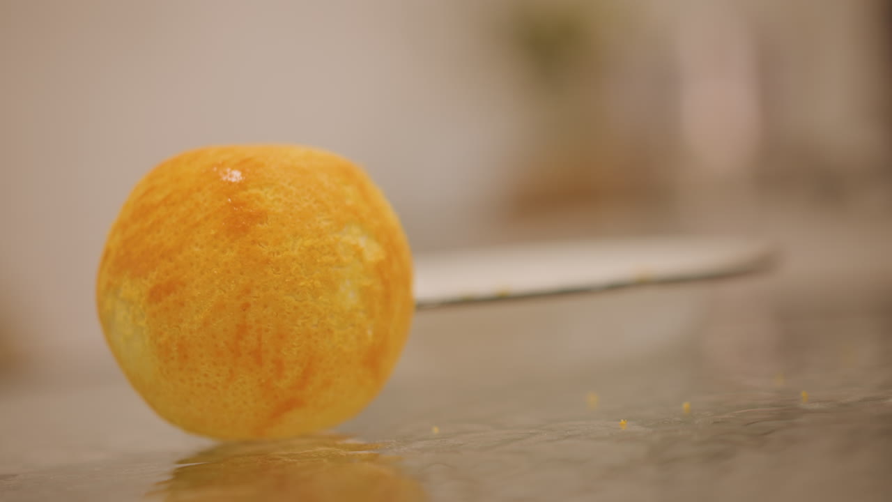 Arancia intera su piano di lavoro in vetro con una mano che si avvicina, buccia brillante e testurizzata con leggera umidità, un delicato rotolamento e un gesto tattile prima di affettare, bokeh caldo della cucina, primo piano sensoriale della preparazione della frutta.