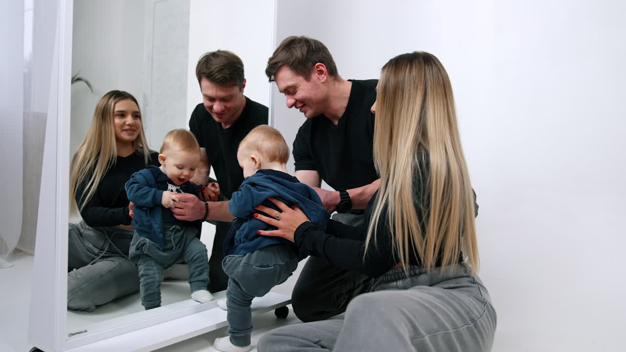 Mom and dad sit near the mirror looking at their baby. Little cute child plays with his reflection.