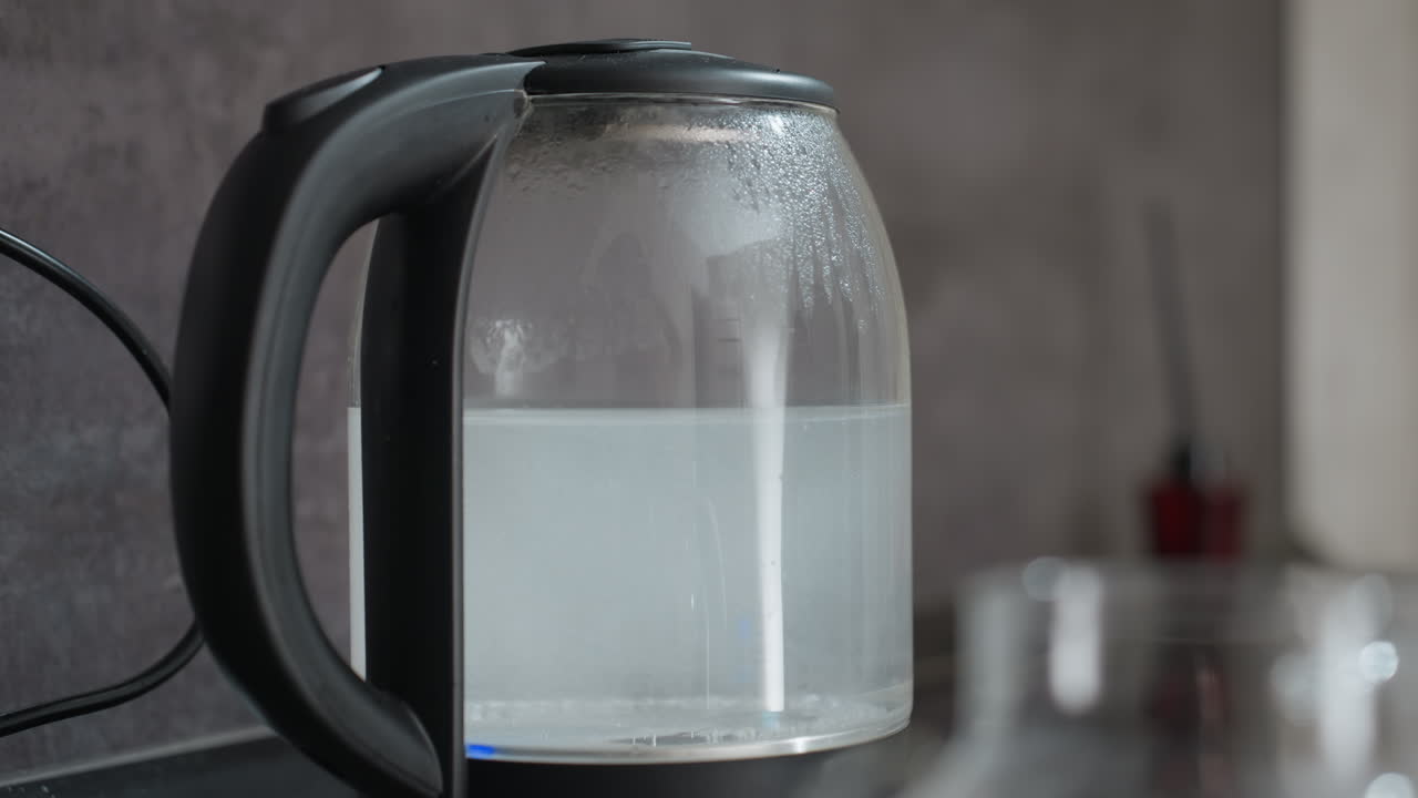 Dampf steigt aus einem blau beleuchteten Kessel auf, eine ruhige Küchenszene mit einem dampfenden Glaskessel mit blauem Licht, eine fokussierte Aufnahme von kochendem Wasser in einem Glaskessel, der inmitten eines gemütlichen Küchenambientes Dampf abgibt